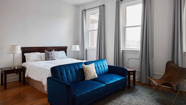 A hotel room with a large bed, a blue sofa, a small table, a chair, and two windows with curtains. - Walkable to Huntington Center, Fifth-Third Field and the Convention Center! (Toledo)