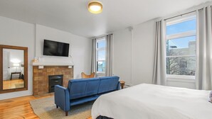 A hotel room with a bed, a blue sofa, a fireplace, a television, and large windows.