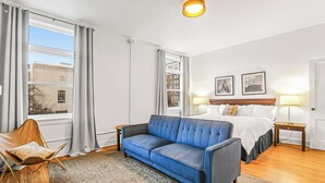Ett hotellrum med en blå soffa, en säng med vita sängkläder, ett trägolv och ett fönster med gardiner.