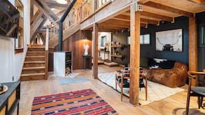 A spacious loft with a high ceiling, exposed wooden beams, a staircase, a cozy seating area with a fur throw, and a large window. - Creekside Cabin with Hot Tub & Wood-Burning Stove (Lanesville)