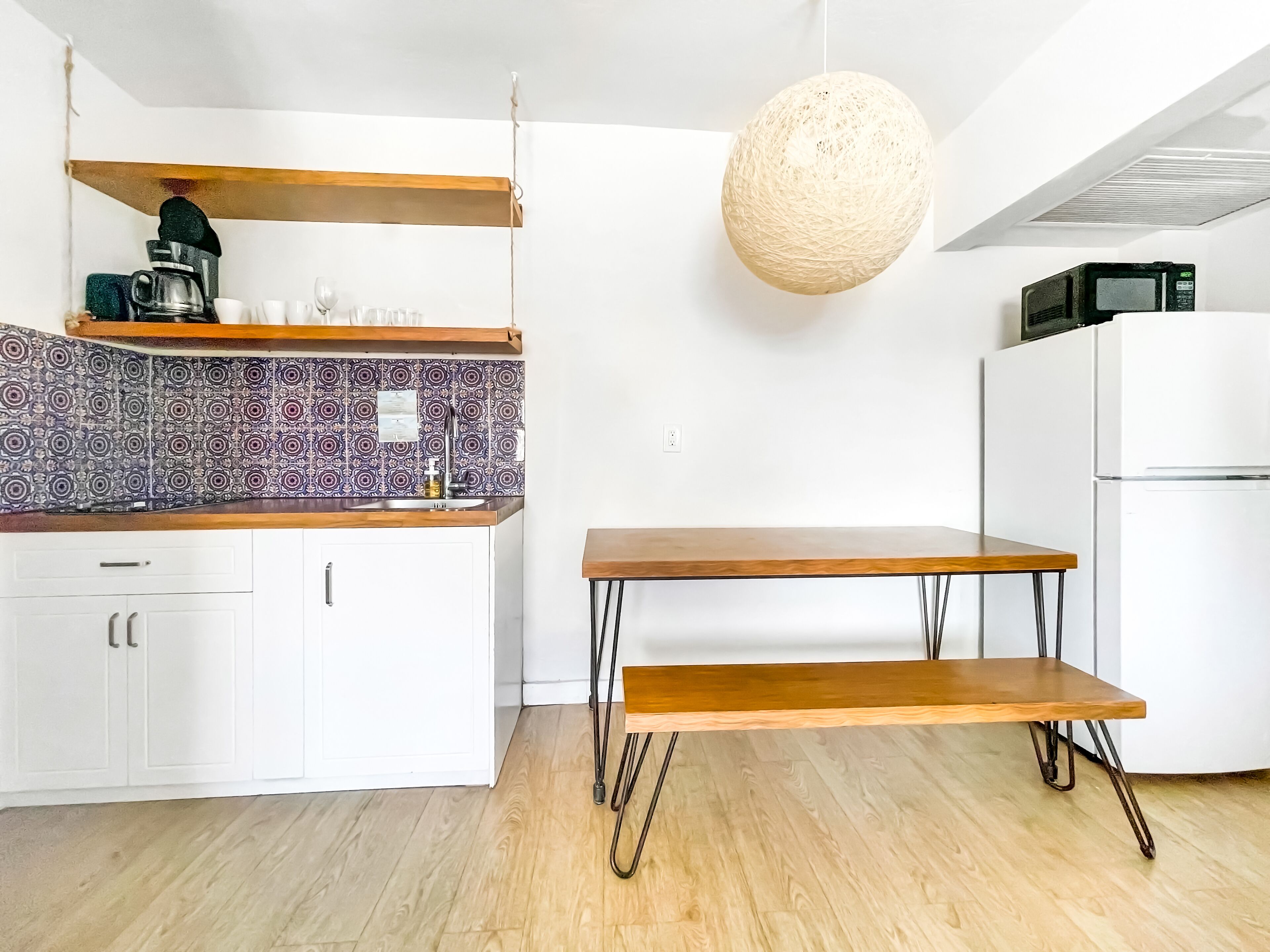 Una cocina moderna con armarios blancos, salpicadero estampado, estantes de madera, una lámpara colgante y una mesa minimalista.