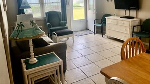 Ein Wohnzimmer mit Sofa, Sessel, kleinem Tisch und Fernseher. Eine Tür führt auf einen Balkon mit Blick ins Freie.