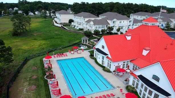 Una piscina grande rodeada de sillas de descanso y sombrillas, con un edificio de techo rojo cerca.
