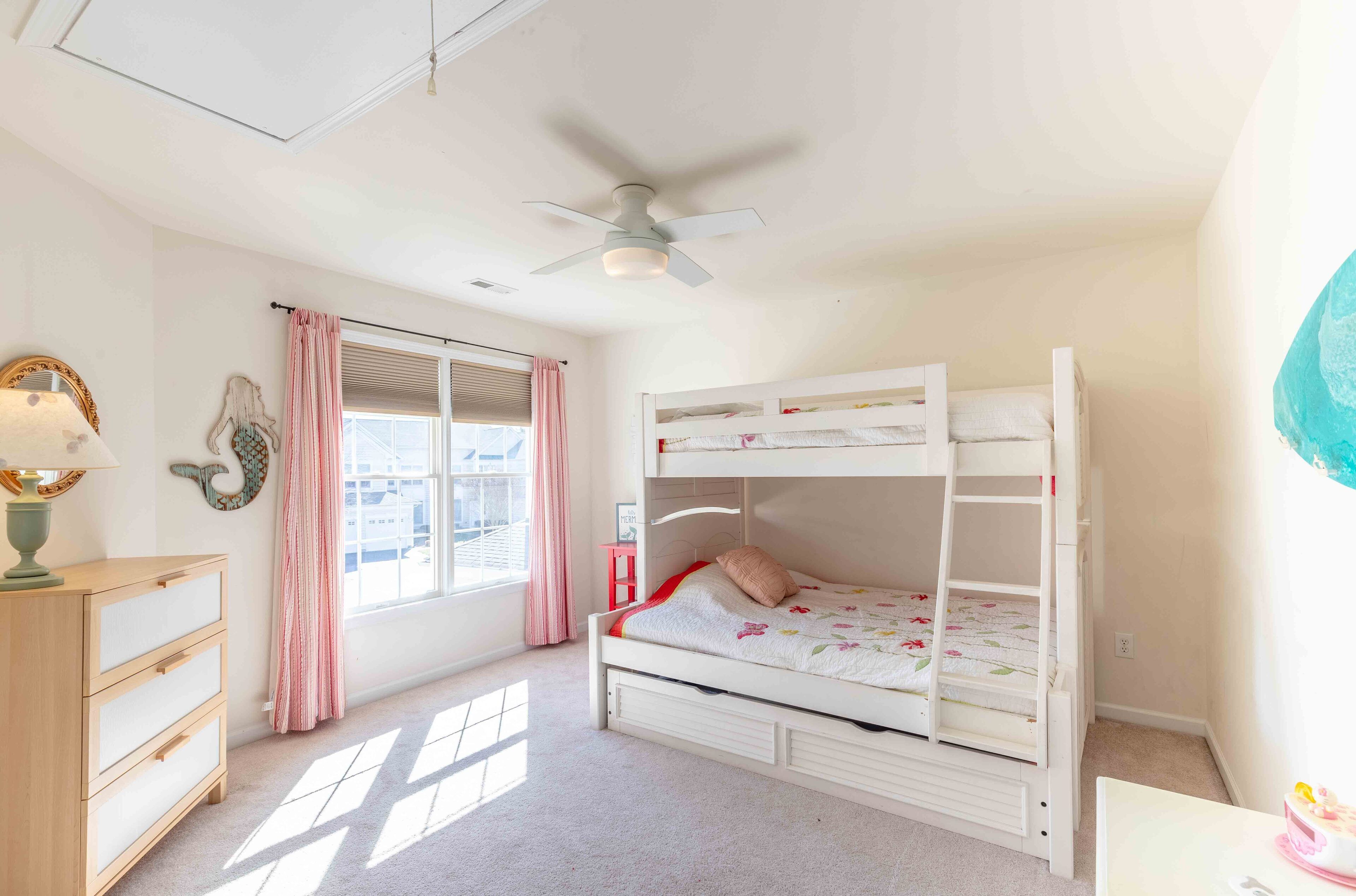 A bunk bed with a ceiling fan, a dresser, a window with curtains, and a wall decoration.