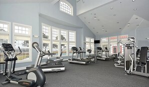 Academia espaçosa com várias esteiras, equipamentos elípticos e estações de musculação. Grandes janelas oferecem iluminação natural e vista para o exterior.