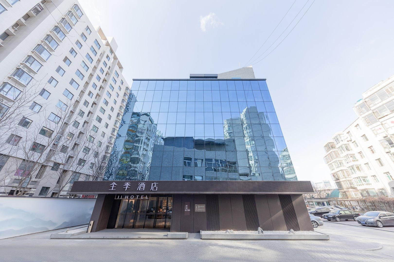 A modern hotel with a glass facade reflecting surrounding buildings, a ground-level entrance, and a visible sign in a foreign language.