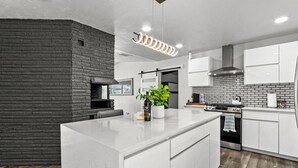 A modern kitchen with white cabinetry, a central island, a brick accent wall, and a stainless steel range hood. - MODERN HOME WITH GYM, SAUNA, POOL AND GAME ROOM (El Paso)