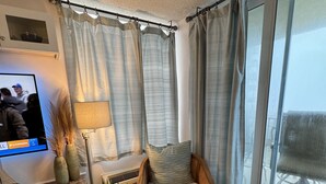 Une chambre d’angle avec un fauteuil, un téléviseur, une plante et une fenêtre avec des rideaux.