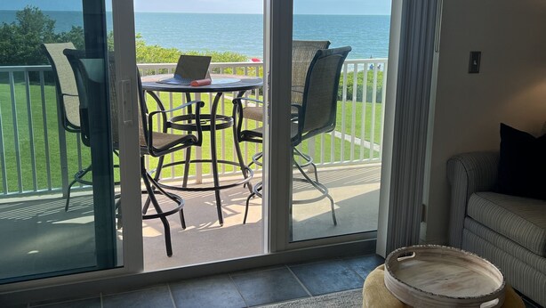 Une pièce dotée d’une porte-fenêtre coulissante donnant sur un balcon avec une table et des chaises, un pouf confortable et une vue sur l’océan.