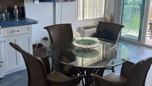 Une cuisine moderne avec une table à manger en verre, des chaises en osier, un téléviseur à écran plat et une vue sur l’extérieur.