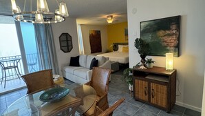 Un coin repas avec une table en verre et des chaises en osier, un salon avec un canapé et un tableau, et un espace cuisine avec un meuble et une plante.