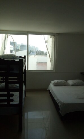 A room with a bunk bed and a single bed, a window with a view of buildings, and a tiled floor.