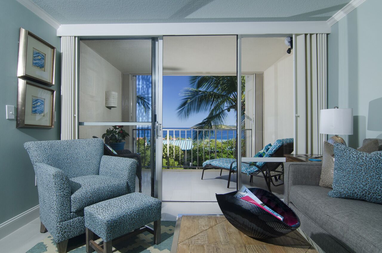 Ein Wohnzimmer mit Sofa, Sessel und einem kleinen Tisch. Eine Glasschiebetür führt zu einem Balkon mit Blick auf Palmen und das Meer.