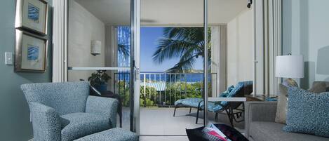 Ein Wohnzimmer mit Sofa, Sessel und einem kleinen Tisch. Eine Glasschiebetür führt zu einem Balkon mit Blick auf Palmen und das Meer.