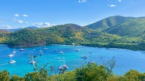 Une baie côtière où de nombreux voiliers sont amarrés dans des eaux d’un bleu limpide, entourée de collines verdoyantes et, au loin, de quelques bâtiments.