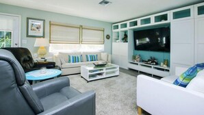 Ein Wohnzimmer mit einem weißen Sofa, einem schwarzen Ledersessel, einem Couchtisch und einem Fernseher auf einer weißen Unterhaltungsanlage.