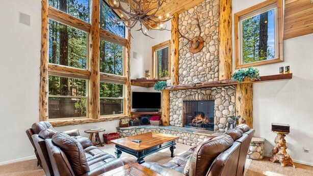 A spacious living room with a stone fireplace, leather sofas, and large windows overlooking a forest. - Snow Summit Bike Ski Estate | Family Friendly (Big Bear Lake)