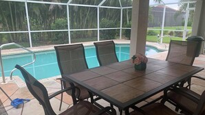 Une terrasse extérieure avec une piscine, entourée de chaises et d’une table, et offrant une vue sur des palmiers et un bâtiment en arrière-plan.