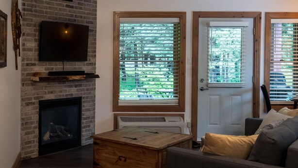A living room with a brick fireplace, a flat-screen TV mounted above it, a wooden coffee table, and a sofa with cushions. There are two windows with blinds, and a door is visible in the background.