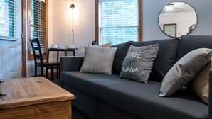 A cozy living room with a dark grey sofa, a wooden coffee table, and a dining table with chairs. There is a large mirror on the wall and a window with blinds.