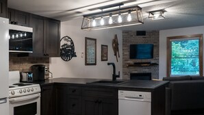 A modern kitchen with a built-in oven, a dishwasher, and a large island with a sink. There is a brick wall and a mounted television above the island.