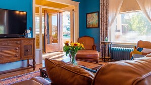 Une pièce avec un mur bleu, une commode en bois, un téléviseur, un canapé en cuir marron, une chaise, une petite table sur laquelle se trouve un vase de fleurs, et une fenêtre avec des rideaux.