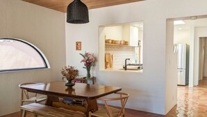 A dining area with a wooden table and chairs, a hanging light fixture, a vase with dried flowers, and a window with a view of the kitchen. - Western Moon - Private Retreat with Pool and Hot Tub (Tucson)