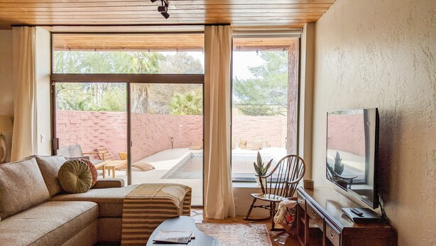 A living room with a sofa, ottoman, coffee table, and two armchairs. There is a television on a wooden cabinet and a sliding glass door leading to an outdoor patio with a brick wall and a pool. - Western Moon - Private Retreat with Pool and Hot Tub (Tucson)