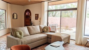 A cozy living room with a sectional sofa, a small table, and a red armchair. There is a large window with a view of a brick wall and greenery outside. - Western Moon - Private Retreat with Pool and Hot Tub (Tucson)