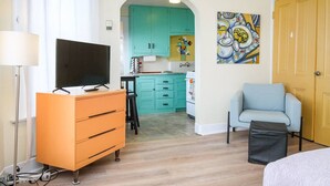 Un espace de vie compact avec une télévision posée sur une commode, une kitchenette dotée de meubles turquoise, un fauteuil bleu clair et une porte ornée d’un tableau décoratif.