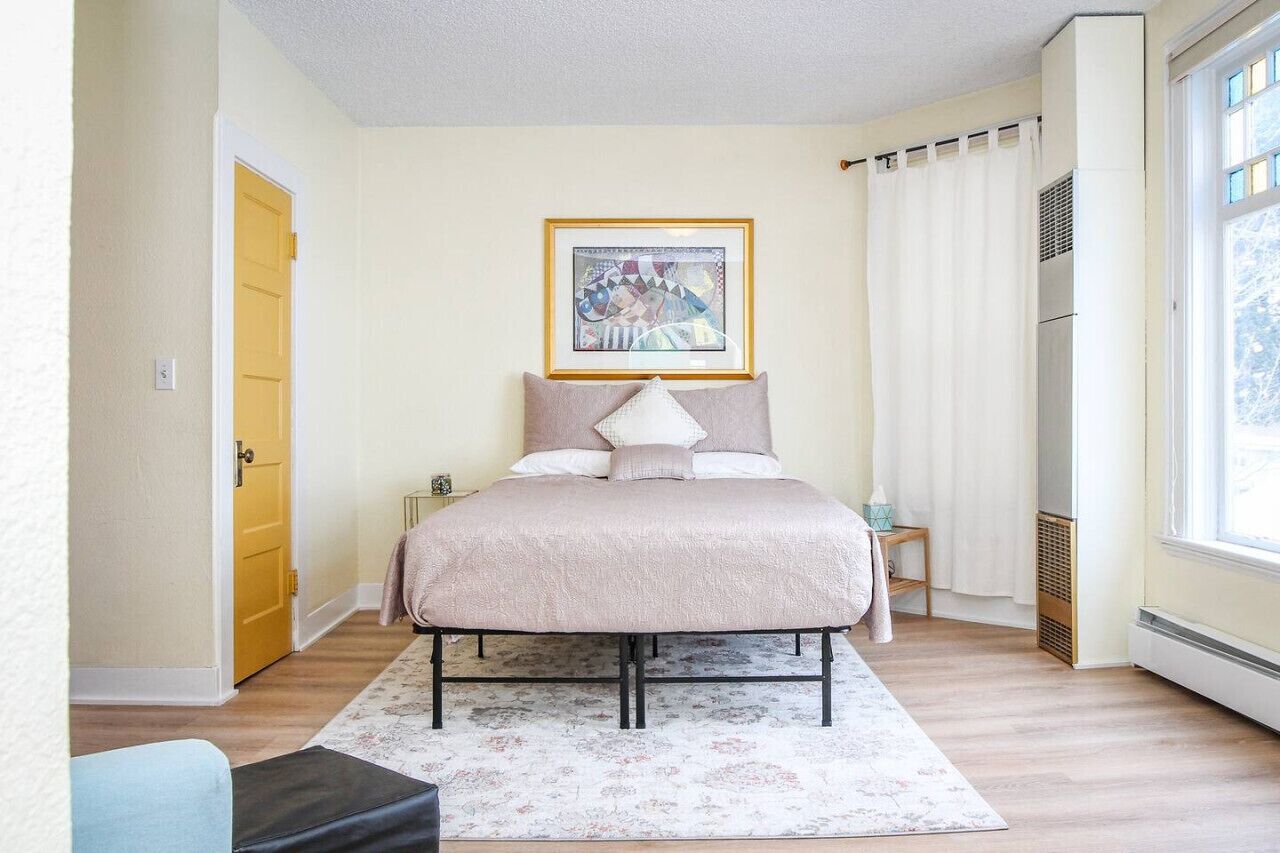 A bedroom with a bed, a framed picture on the wall, a window with curtains, and a radiator.