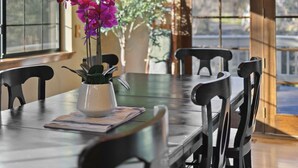 Uma sala de jantar com teto de madeira, um candelabro, uma mesa com cadeiras e um vaso com flores roxas.