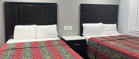 A hotel room with two beds, each with a black headboard, white bedding, and a red and green patterned bedspread. There is a small black nightstand between the beds.