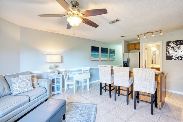 A modern living room with a ceiling fan, a dining area with white chairs, and a kitchen visible in the background. - walk to the beach 1 minute across (Miramar Beach)