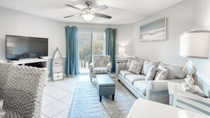 Ein modernes Wohnzimmer mit einem Flachbildfernseher, Deckenventilator, einer Couch, einem Couchtisch und einem großen Fenster mit Vorhängen.