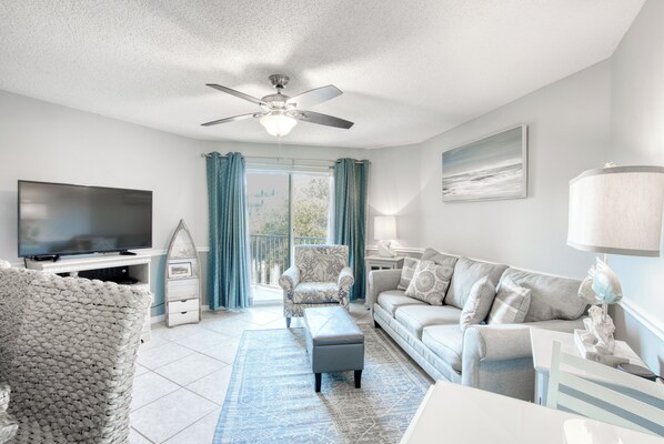 A modern living room with a flat-screen TV, a ceiling fan, a sofa, a coffee table, and a large window with curtains.