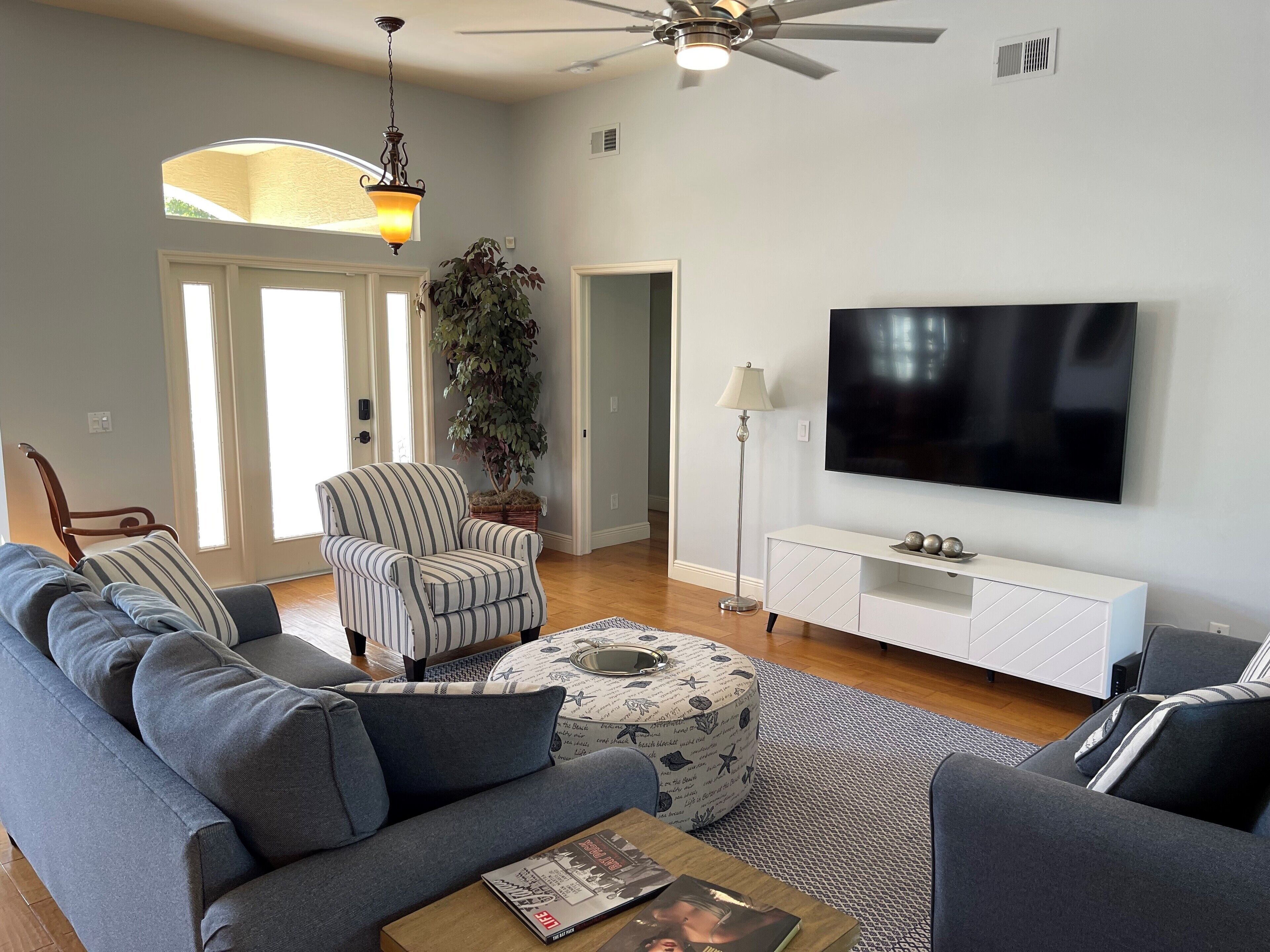 A living room with a grey sofa, striped armchair, ottoman, and a flat-screen TV on a white cabinet.