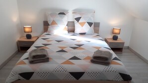 A neatly made bed with a geometric patterned bedspread, two pillows, and two folded towels on the side. There are two bedside tables with lamps and a nightstand with a drawer.