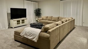 Una sala moderna con un sofá seccional color beige, un televisor de pantalla plana sobre un mueble blanco, y una mesa de centro redonda con un objeto decorativo.