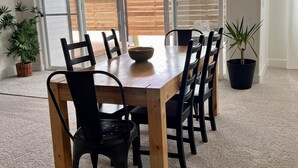 Un comedor con una mesa y sillas de madera, una planta en maceta y vistas a una terraza de madera al aire libre a través de puertas de vidrio grandes.