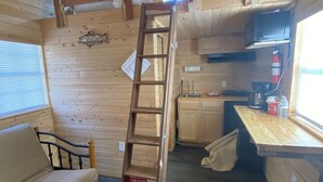 A cabin interior with a wooden ladder, a sofa, a kitchen area, and a window with blinds.