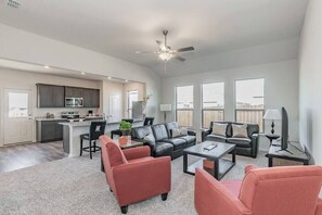 A spacious living room with a large window, a ceiling fan, and a kitchen area with dark cabinets and modern appliances.