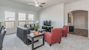 Ein modernes Wohnzimmer mit einem grauen Sofa, roten Sessel, einem Couchtisch und einem Fernseher. Es gibt große Fenster und einen Deckenventilator.