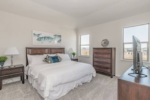 A bedroom with a large bed, a nightstand with a lamp, a dresser, and a TV on a desk. - Modern Spacious 4BR New Built Retreat ~ Fam&Group (Seagoville)