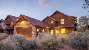 Une maison rustique à deux étages, avec des fondations en pierre et un revêtement extérieur en bois, entourée d’une végétation aride.