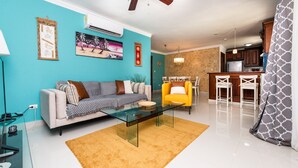 A modern living room with a glass coffee table, a sofa, and a yellow armchair. There is a kitchen area with a dining table and bar stools. - Recently refurbished, five star facility (El Cortiesito-)