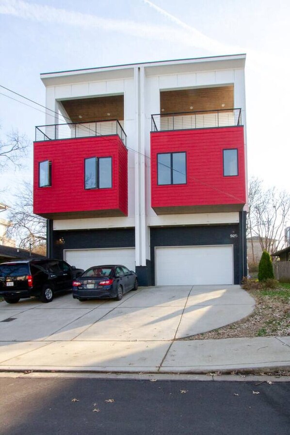 Un edificio residencial moderno de dos pisos con fachada roja, dos puertas de garaje y un estacionamiento con dos coches.