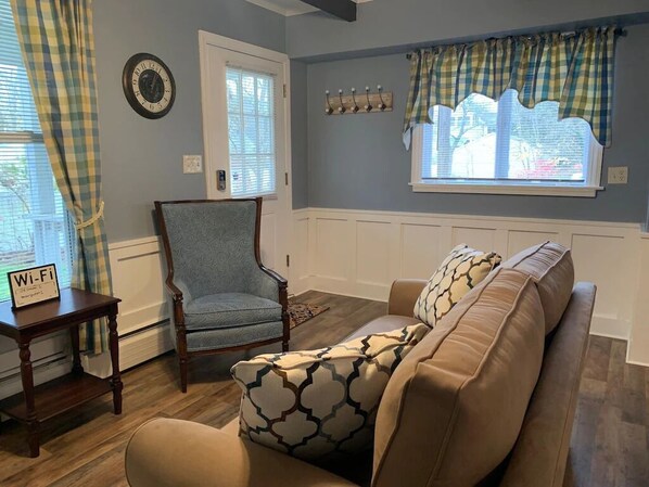 Ein Wohnzimmer mit einem beigefarbenen Sofa, einem blauen Sessel, einem Holztischchen mit einem Wi-Fi-Symbol, einer Wanduhr und einem Fenster mit Karottenvorhängen.