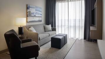 A modern living room with a grey sofa, a black ottoman, a side table with a lamp, and a flat-screen TV mounted on the wall.