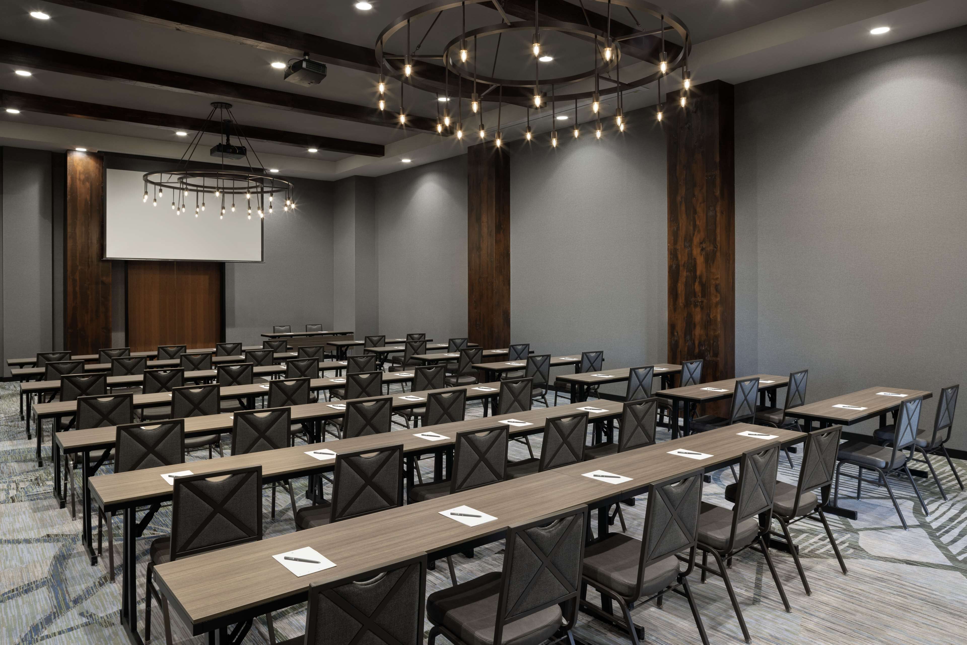 Un salón de conferencias con mesas y sillas dispuestas en filas, una pantalla grande y una moderna lámpara de araña.